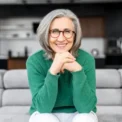 Healthy aged woman with grey hair with glowing blue eyes, elegant glasses, sitting on the pure comfortable sofa at modern apartment, dressed in green sweater and white pants, put chin on the hands