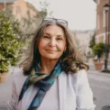 Outdoor image of gorgeous positive lady with charming smile and loose gray hair enjoying nice summer day, wearing white jacket, silk scarf and eyeglasses. Beauty, urban style and fashion concept